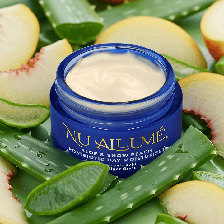 An open jar of Nu Allumé Aloe and Snow Peach Postbiotic Day Moisturiser surrounded by cut pieces of aloe vera and snow peaches