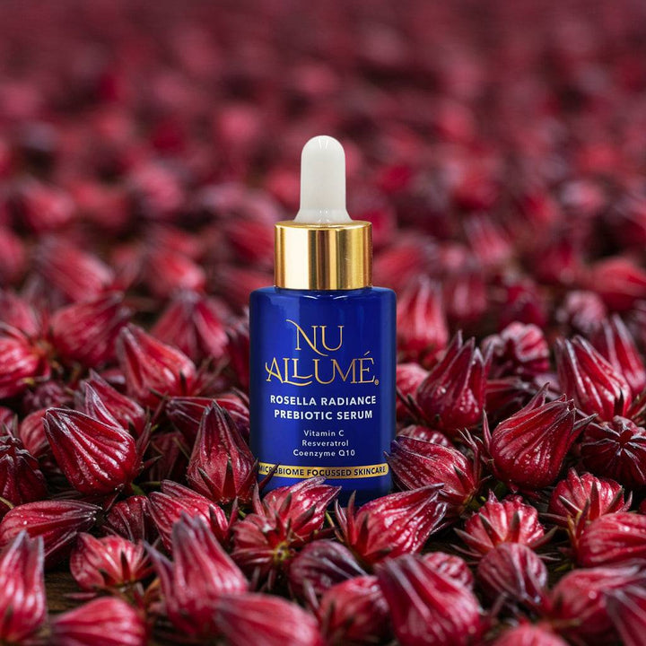 A bottle of Nu Allumé Rosella Radiance Prebiotic Serum Surrounded by Rosella Buds